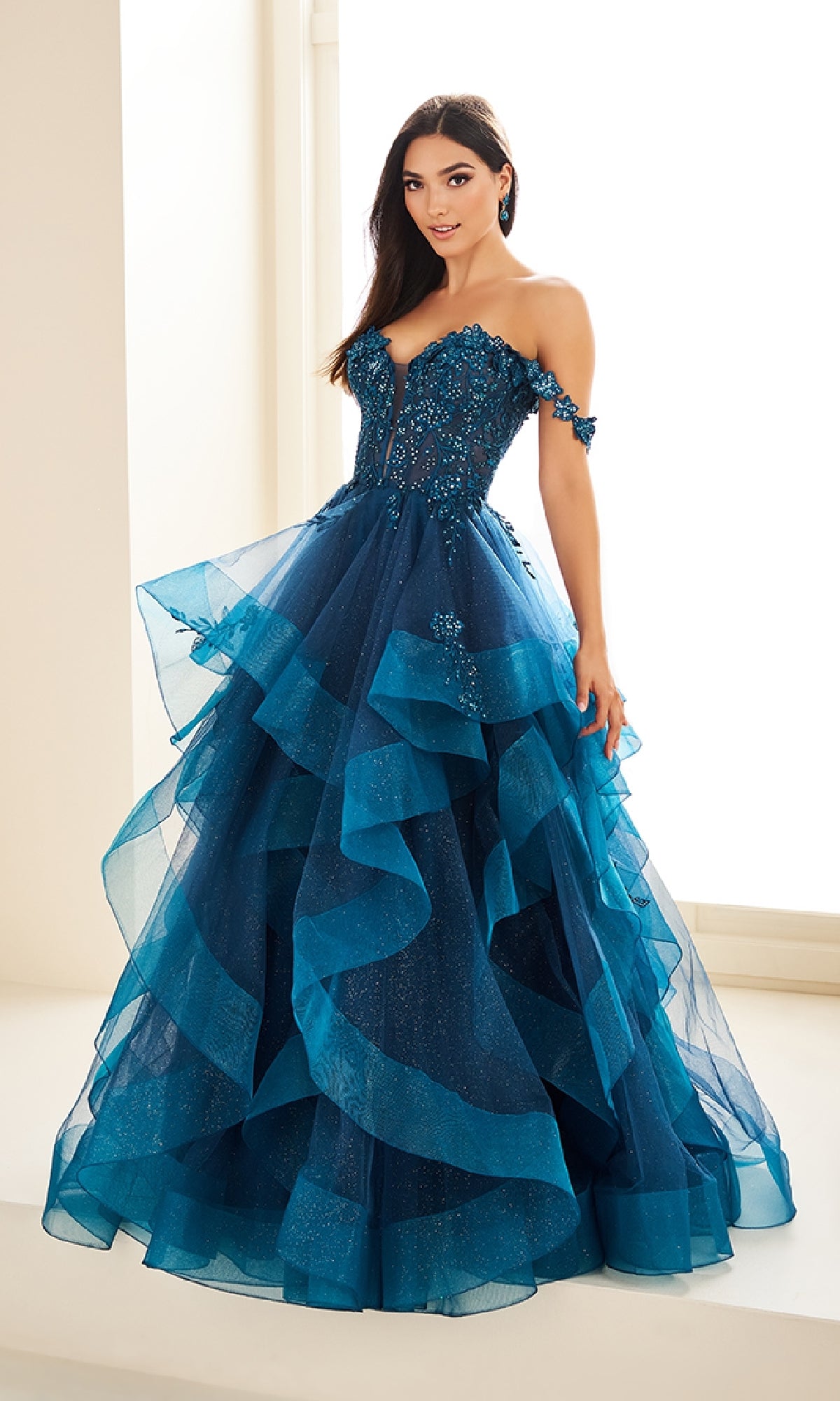 A woman stands indoors near a window wearing the Ellie Wilde Long Designer Prom Ball Gown EW35084—a blue off-the-shoulder evening dress with a tiered ruffled skirt, floral embellishments, and glitter tulle.